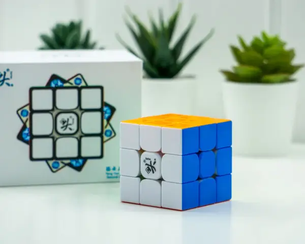 En speed cube 3x3 med orange, vit och blå sida står bredvid sin förpackning. Tre gröna växter i vita krukor syns i bakgrunden.