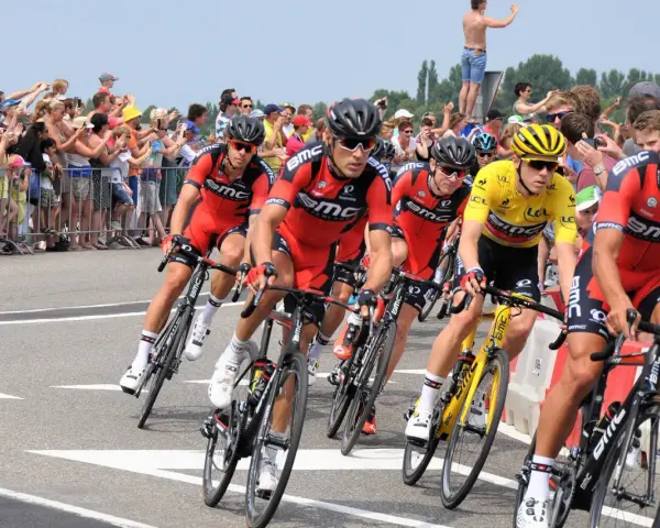 Cyclists in red BMC jerseys race, with one in yellow, as spectators cheer. A perfect scene to watch Tour de France live!