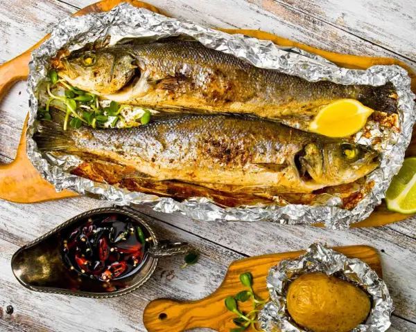 Due branzini cotti al cartoccio, dorati e succulenti, serviti con spicchi di limone, patata al cartoccio e salsa piccante.