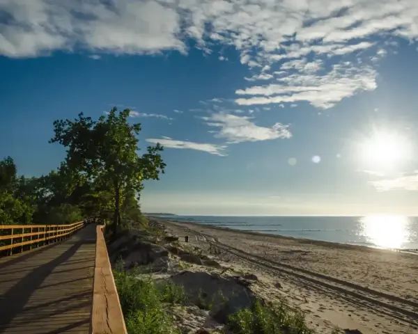 Drewniana ścieżka wzdłuż plaży, idealna na przejażdżkę rowerem po trasie rowerowej R10. Słońce odbija się w morzu.