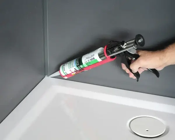 Dłoń trzyma pistolet z tubą **sylikonu**, aplikując go w rogu brodzika prysznicowego.
