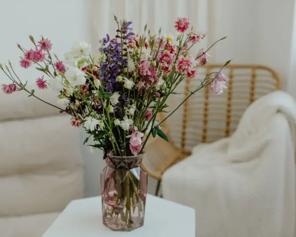 Wazon z kolorowymi kwiatami ciętymi, wśród nich różowe dzwonki i białe margaretki, stoi na stoliku obok plecionej wiklinowej fotel.