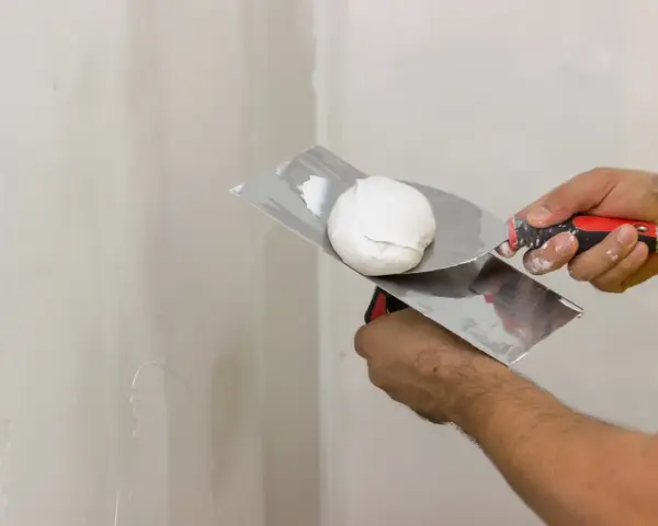 Mani che usano spatole per applicare stucco su un muro. Lo stucco serve a livellare e riparare le imperfezioni.