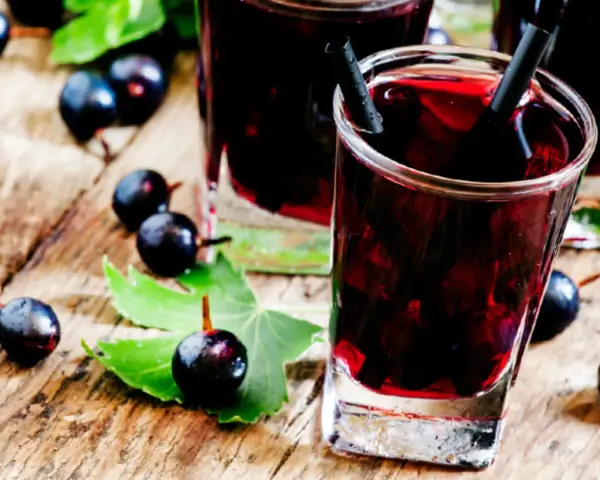 Gläser mit dunklem Johannisbeeren Schnaps, umgeben von frischen Beeren und Blättern auf Holz.