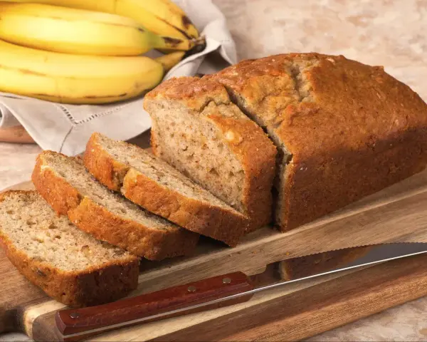 Pyszny, domowy chlebek bananowy, pokrojony na kromki, leży obok dojrzałych bananów na desce.