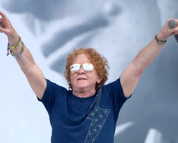 Wokalista Simply Red z uniesionymi rękami, trzymający mikrofon. Ma charakterystyczne rude włosy i okulary przeciwsłoneczne.
