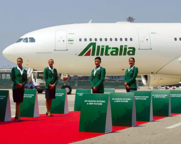 Cztery stewardessy w zielonych uniformach stoją na czerwonym dywanie przed samolotem Alitalia.