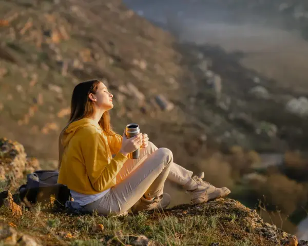 A young woman enjoys the art of doing nothing, holding a thermos and basking in the sun on a rocky hillside.