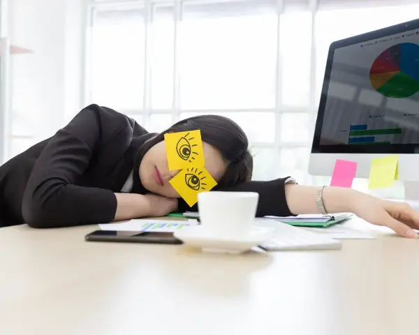 En kvinna sover vid sitt skrivbord med post-it-lappar som föreställer ögon på ögonlocken. Det är uppenbart att hon tycker det är tråkigt på jobbet.