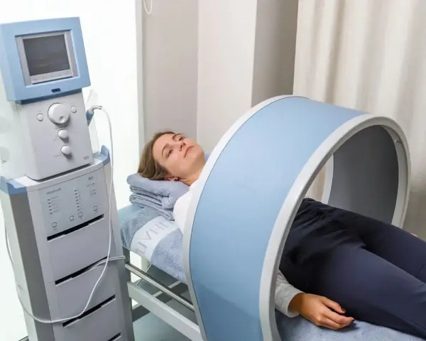 Kobieta leży na łóżku rehabilitacyjnym, poddając się zabiegowi z wykorzystaniem urządzenia generującego pole magnetyczne, które obejmuje jej kolano.