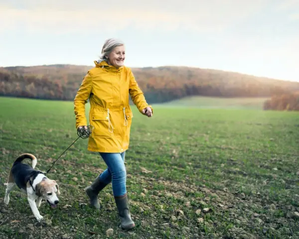Glad kvinna i gul jacka njuter av en promenad med sin hund i ett fält. En härlig promenad som bränner kalorier.