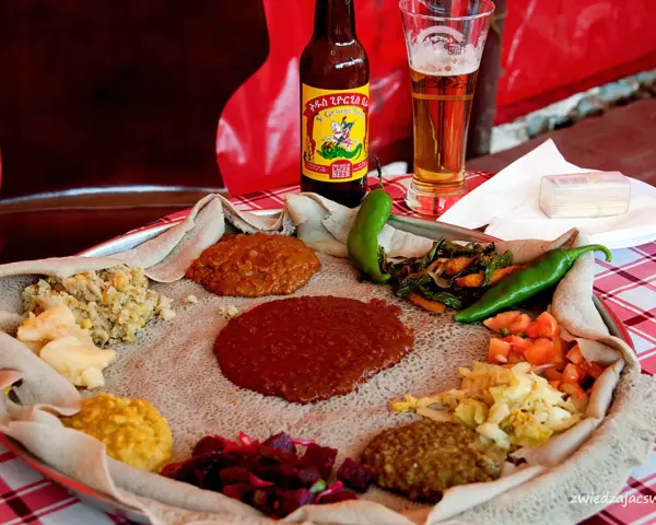 Tradycyjna kuchnia etiopska na płaskim chlebie injera, z różnymi gulaszami, warzywami i papryczkami. Obok piwo.
