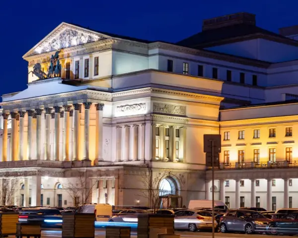 Teatr Wielki w Warszawie, najstarszy teatr w Polsce, nocą. Monumentalna fasada z kolumnami i rzeźbami, oświetlona ciepłym światłem, z samochodami w ruchu ulicznym przed budynkiem.