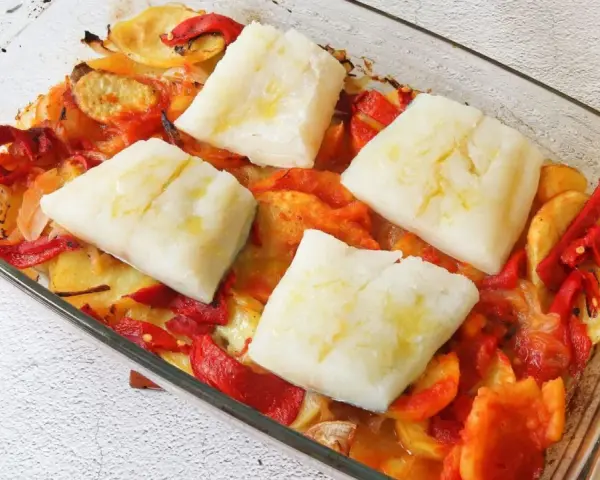 Cuatro porciones de bacalao al horno reposan sobre patatas y pimientos asados en una fuente de vidrio.