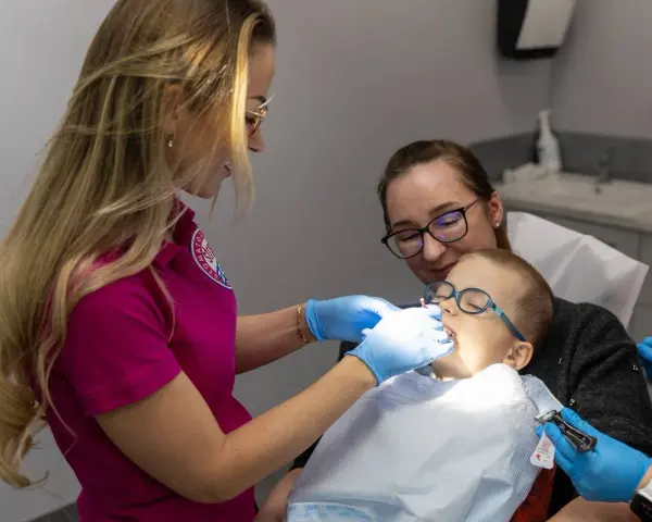 Dentystka bada zęby chłopca, podczas gdy jego mama go wspiera. Zwracają uwagę na zdrowie dziąseł, by zapobiec recesji dziąseł.