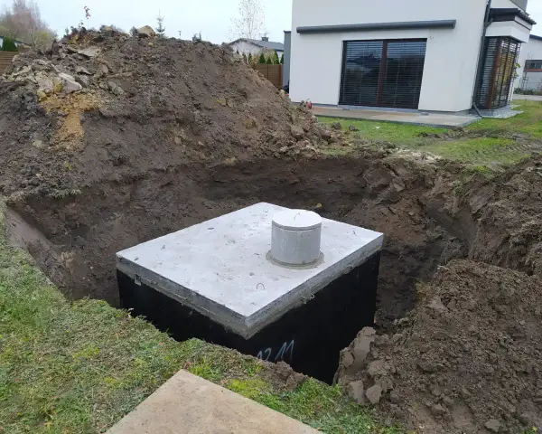 Gotowy odpływ z rynny do ziemi, betonowy zbiornik w wykopie, obok hałda ziemi i nowoczesny dom.