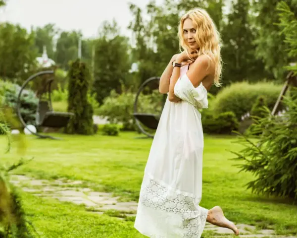 Uśmiechnięta kobieta panna w zwiewnej białej sukni, z rozpuszczonymi blond włosami, stoi na trawniku w ogrodzie.
