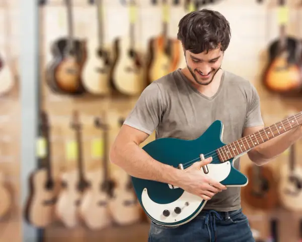 Uśmiechnięty mężczyzna testuje gitarę elektryczną w sklepie muzycznym. W tle widać wiele innych instrumentów.
