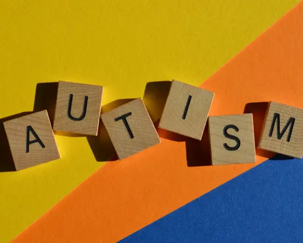Lettere di legno formano la parola AUTISM su uno sfondo colorato. La domanda "autistici si nasce o si diventa" risuona.