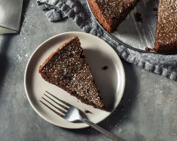 Una fetta di torta all'acqua al cacao, spolverata di zucchero a velo, su un piatto con una forchetta. Accanto, la torta intera e una paletta.