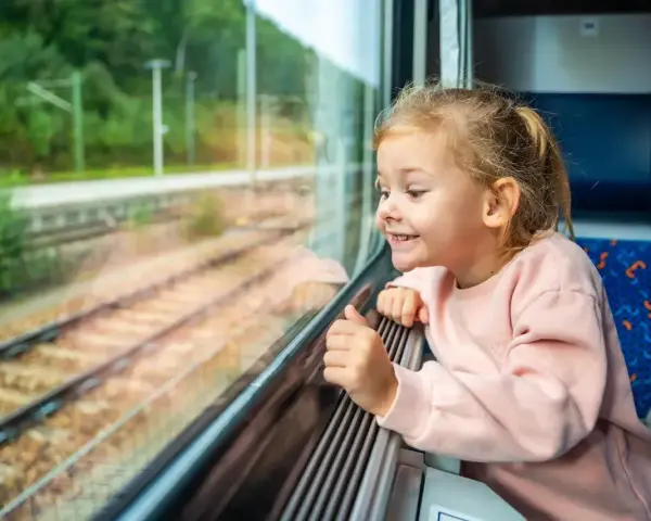 Uśmiechnięta dziewczynka patrzy przez okno pociągu. Zastanawia się, od ilu lat można jeździć samemu pociągiem, marząc o podróżach.