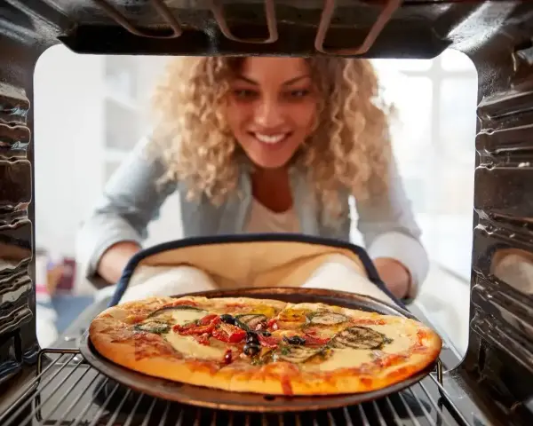 Uśmiechnięta kobieta wyjmuje z piekarnika pyszną, gorącą pizzę. Właśnie skończyła piec pizzę i jest gotowa do jedzenia.