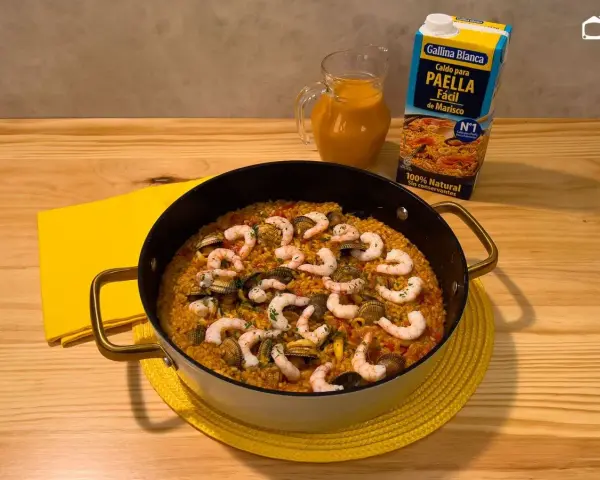 Delicioso arroz a la marinera en paellera, con gambas y mejillones. Al lado, un cartón de caldo para paella de marisco y una jarra con líquido naranja.