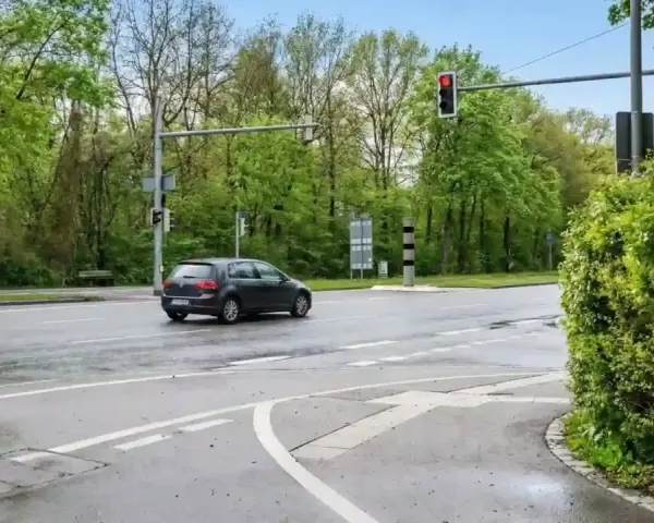 Ein dunkles Auto fährt auf nasser Straße an einer roten Ampel vorbei, neben der ein ampel blitzer steht.
