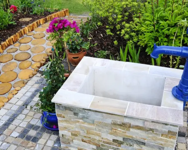 Ein Gartenweg führt zu einem frisch gebauten Brunnen mit blauer Handpumpe.