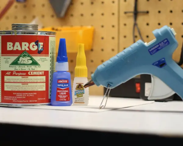 A hot glue gun, Barge cement, Loctite super glue, and Gorilla Super Glue are on a workbench. Does Gorilla Glue melt? This image shows various adhesives.
