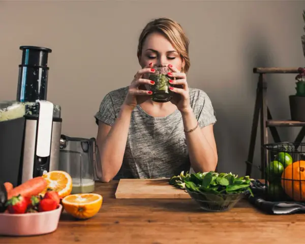 Kvinna dricker grön juice, omgiven av färsk frukt och grönsaker. Hon verkar njuta av sin hälsosamma dryck, kanske som en del av hur deffar man.