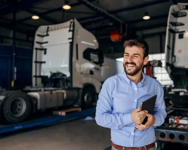Uśmiechnięty mężczyzna z tabletem w ręku, w tle warsztat z ciężarówkami. Oferuje **usługę transportową ryczałt**.