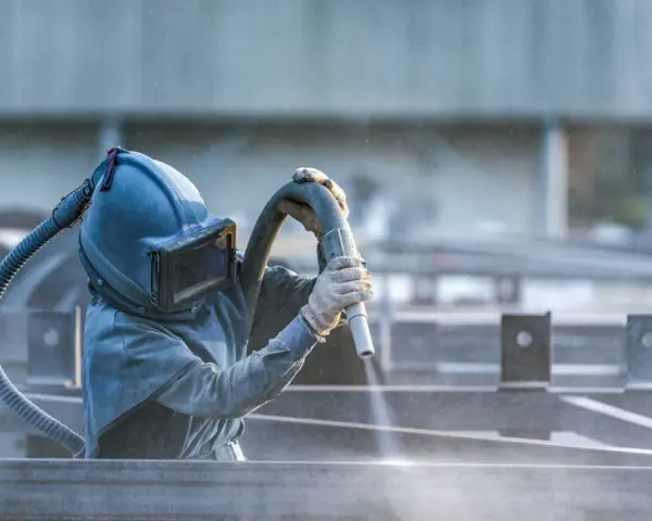 Pracownik w masce piaskuje metalowe elementy. To część pracy z maszynami do metalu, gdzie precyzja jest kluczowa.