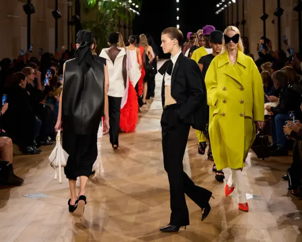 Sfilata Balenciaga: modelli con abiti audaci, tra cui un cappotto giallo acceso e un completo nero, sotto gli occhi del pubblico.