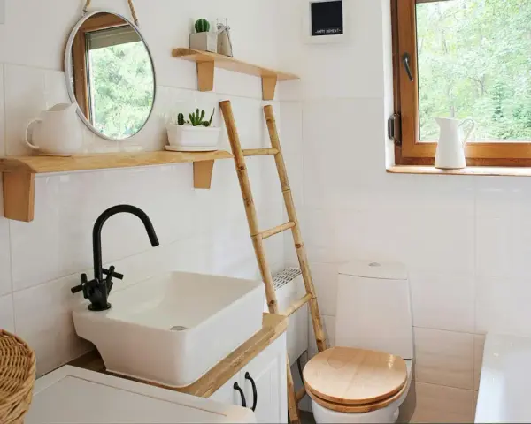 Bagno piccolo con piastrelle bianche lucide, lavabo squadrato, specchio rotondo e scala in bambù. Idee piastrelle bagno piccolo: la luminosità è la chiave.