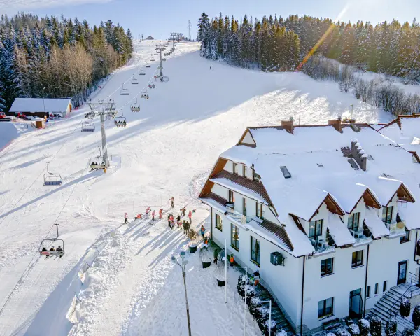 Hotel ze stokiem narciarskim dla dzieci. Kolejka krzesełkowa wwozi narciarzy na ośnieżony stok, a obok hotelu bawią się dzieci.