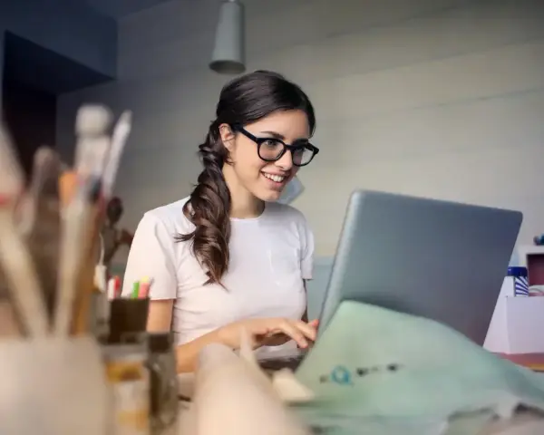 Uśmiechnięta kobieta w okularach pracuje przy laptopie, analizując koszty pracodawcy związane z umową zlecenie.