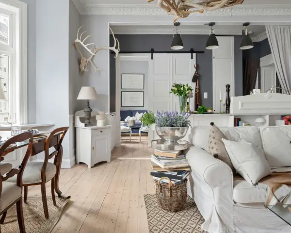 Un salotto con un'atmosfera da arredamento francese chic. Tavolo da pranzo in legno, divano bianco accogliente, lampade e decorazioni rustiche.