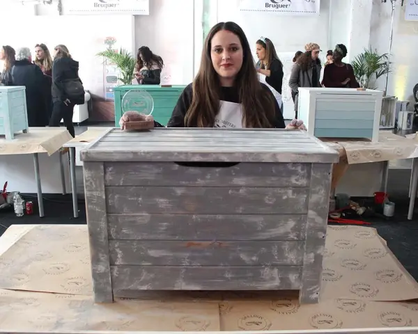 Una mujer muestra un baúl pintado a la tiza con un acabado desgastado. Otros baúles decorados se ven en el fondo.