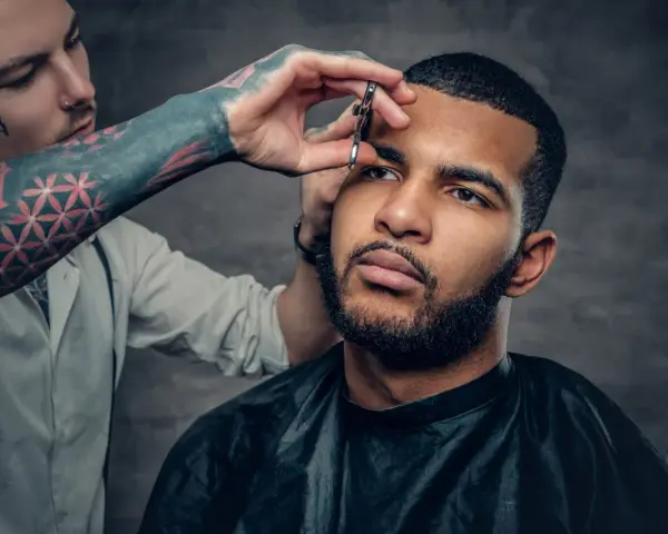 Barbero con tatuajes en el brazo recorta las cejas de un hombre con barba, creando un look buzz cut.