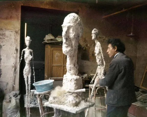 Alberto Giacometti w pracowni, otoczony swoimi charakterystycznymi, smukłymi rzeźbami i niedokończonymi formami.