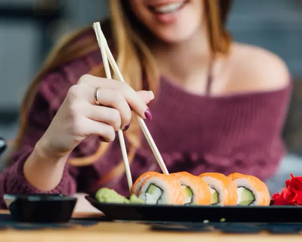 Uśmiechnięta kobieta w ciąży sięga po sushi pałeczkami. Na talerzu łosoś, awokado i imbir.