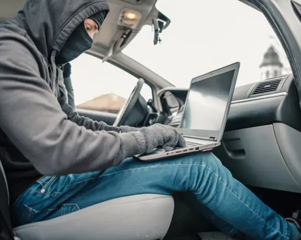Haker w masce i kapturze próbuje obejść zabezpieczenia keyless go, używając laptopa w samochodzie.