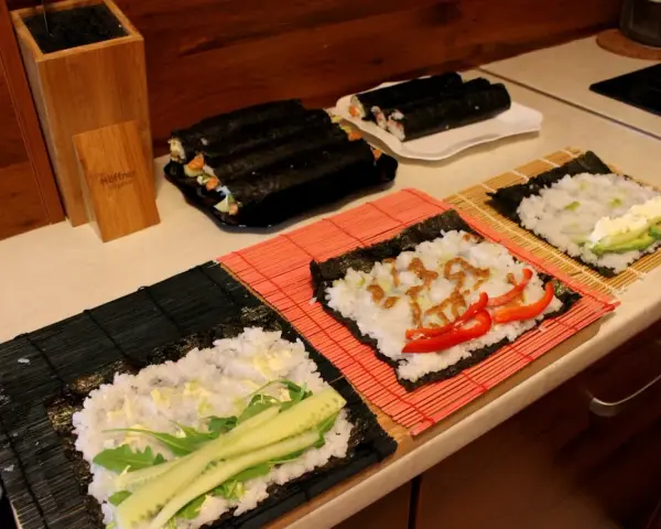 Przygotowanie ryżu do sushi. Na matach leżą arkusze nori z ryżem i dodatkami: ogórkiem, awokado, papryką i sosem.