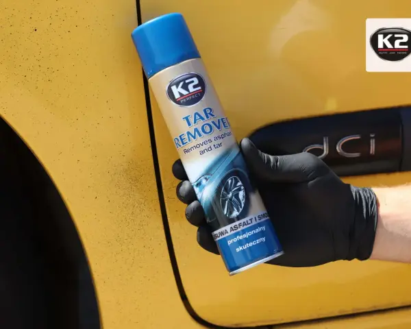 Rękawiczka z czarną gumą trzyma puszkę K2 Tar Remover, która skutecznie usuwa smołę z żółtego lakieru samochodu.