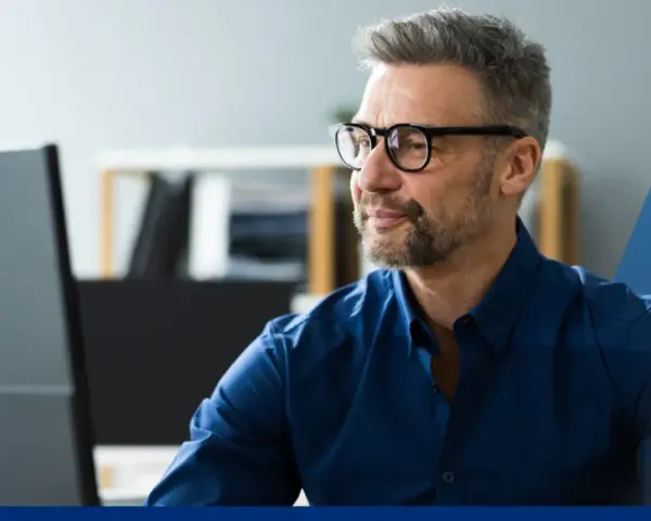 Hombre con gafas y barba gris, concentrado frente a su ordenador. Su camisa azul oscuro es un ejemplo de **art 16 et** en el vestuario profesional.
