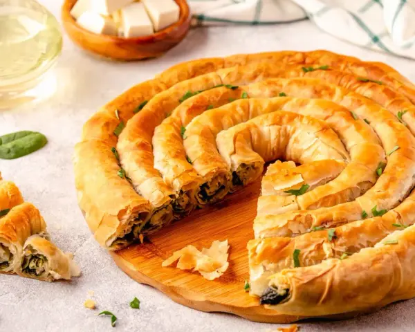 Złocisty borek ze szpinakiem, zwinięty w spiralę, kusi chrupiącym ciastem i aromatycznym nadzieniem. Obok kilka porcji i świeże liście szpinaku.