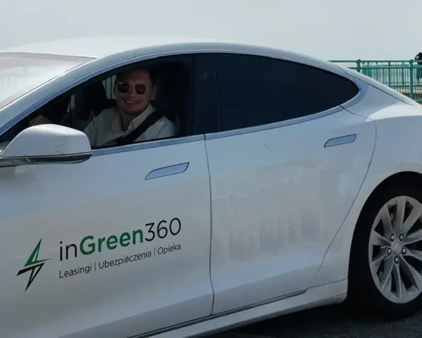 Uśmiechnięty mężczyzna w okularach przeciwsłonecznych siedzi za kierownicą białej Tesli z logo inGreen360.