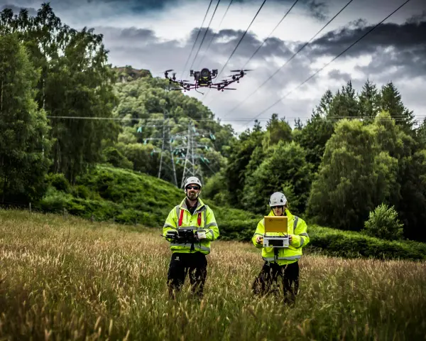 Two technicians in high-visibility jackets operate a power line drone in a grassy field, with a transmission tower and trees in the background.