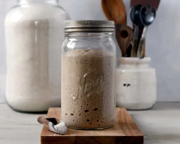 Blubbernde Sauerteig Kultur in einem Glas, bereit für das nächste Brot. Daneben ein Löffel mit Mehl und ein großer Behälter mit weiteren Zutaten.
